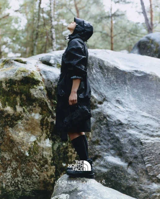 A woman in a black hooded jacket and boots stands on rocks in a forest, looking up, with the The North Face logo at the bottom.