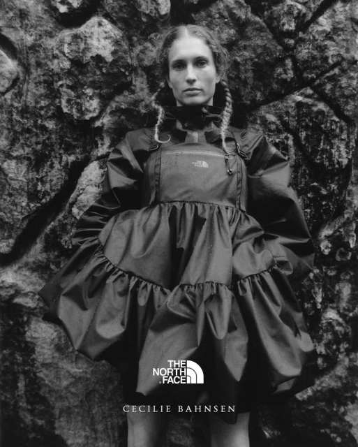 A woman with braided hair wearing a layered black dress stands against a textured gray rock background in a fashion editorial shot.