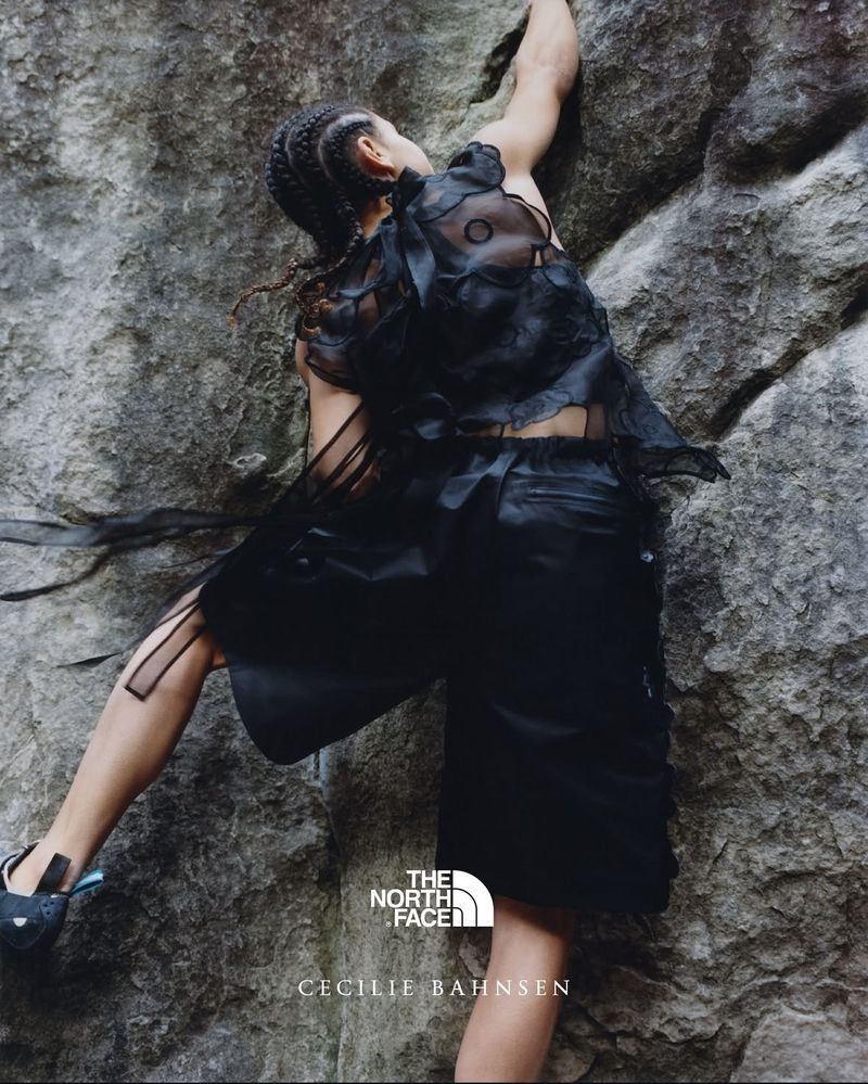 A woman in a black outfit rock climbing outdoors on a gray rock face, showcasing fashion from The North Face and Cecilie Bahnsen.