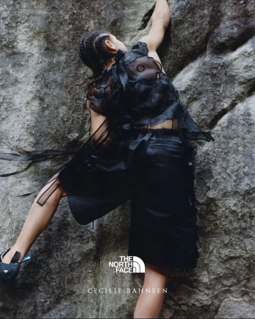 A woman in a black outfit rock climbing outdoors on a gray rock face, showcasing fashion from The North Face and Cecilie Bahnsen.