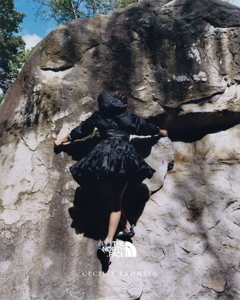 A woman in a dark dress and jacket scaling a large rock face, shot from a low angle with a glimpse of the sky and trees.