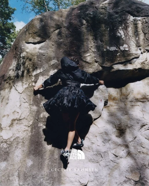 A woman in a dark dress and jacket scaling a large rock face, shot from a low angle with a glimpse of the sky and trees.