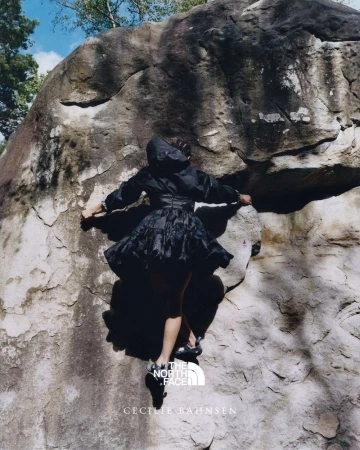 A woman in a dark dress and jacket scaling a large rock face, shot from a low angle with a glimpse of the sky and trees.