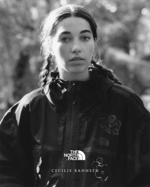 Black and white portrait of a woman in a dark jacket with braids, against a blurry outdoor background with trees and foliage.