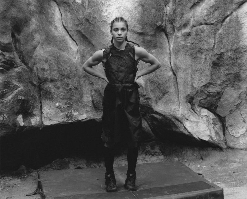 Black and white portrait of a muscular woman standing in athletic apparel, posing in front of a textured rock wall on a gray mat.