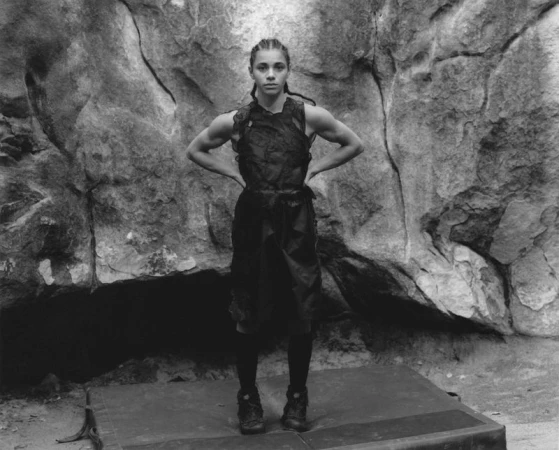 Black and white portrait of a muscular woman standing in athletic apparel, posing in front of a textured rock wall on a gray mat.