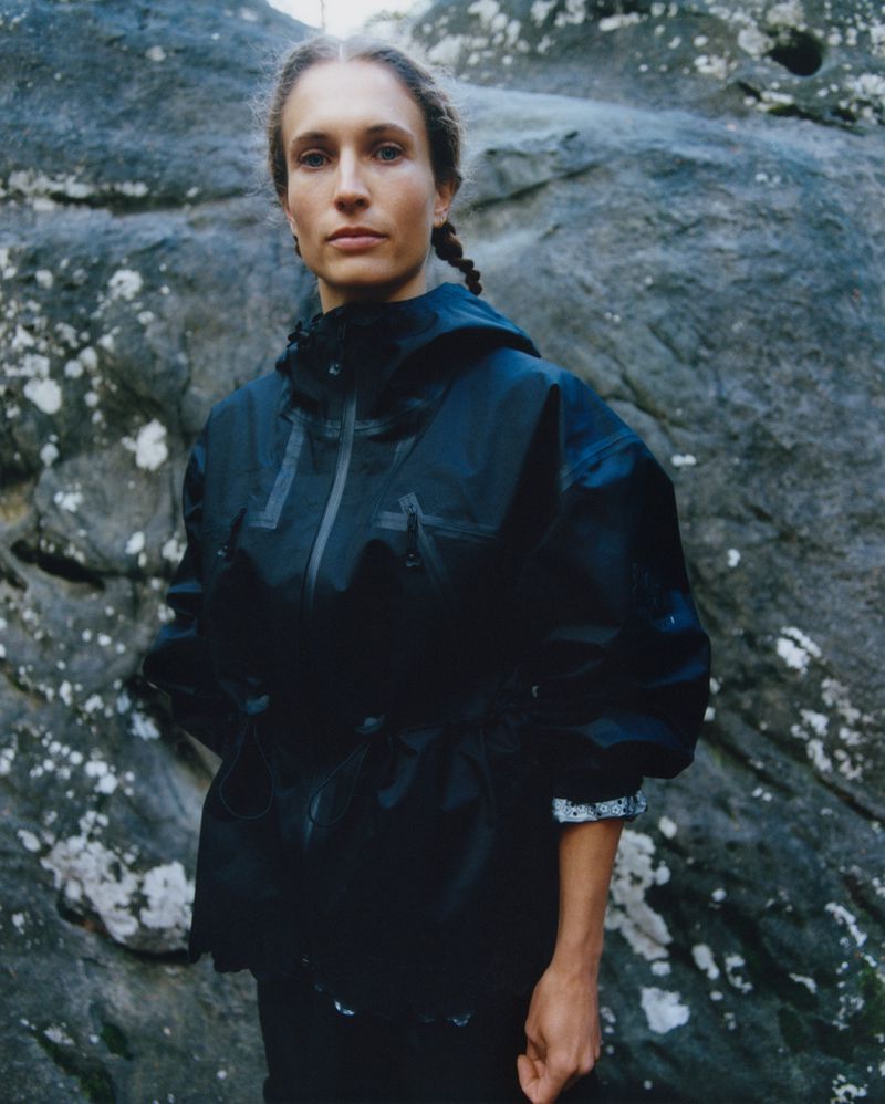 Portrait of a woman wearing a black hooded jacket posing in front of a grey boulder with lichen on an overcast day.