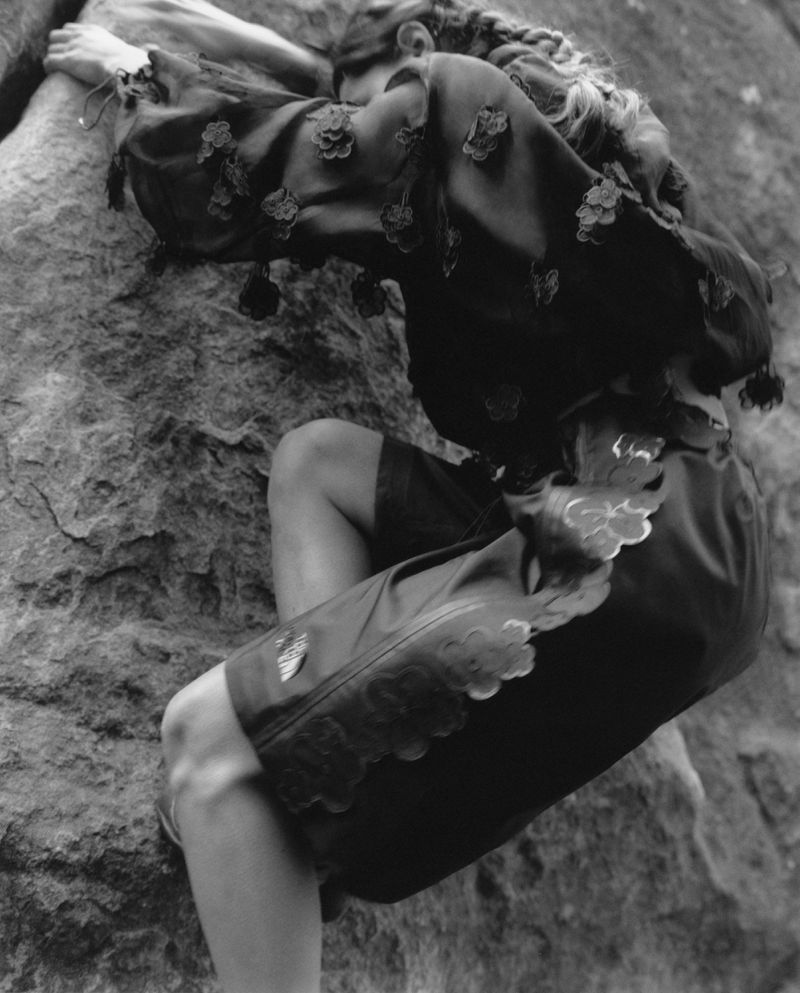 Monochromatic shot of a woman in decorative top and shorts clinging to a textured rock face, showcasing climbing or a unique fashion shoot.