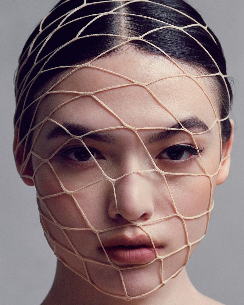 Close-up portrait of an Asian woman with dark hair, light skin, wearing a beige net over her face, set against a neutral gray background.