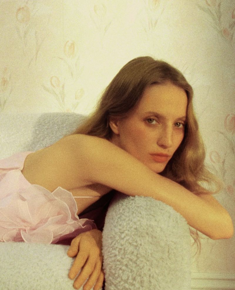 A fair-skinned woman with long hair lies on a blue sofa in a pink dress in an indoor setting with floral wallpaper.