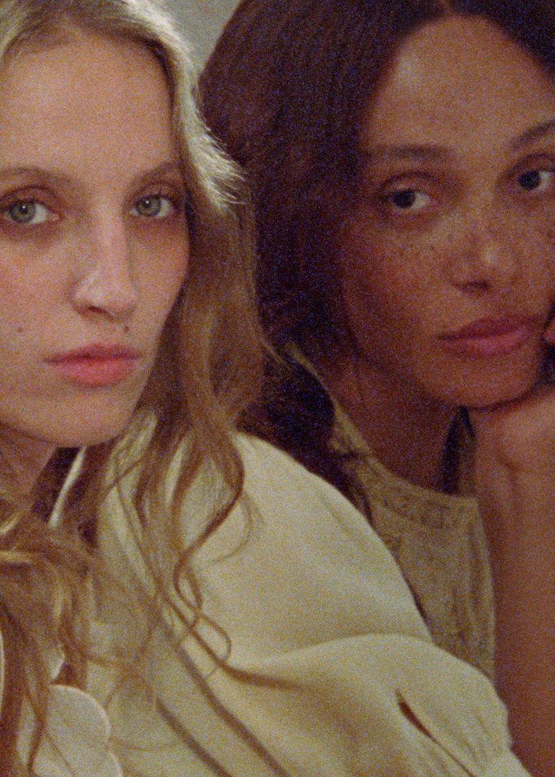 Close-up of two women with fair skin, light hair, and dark hair wearing light colored tops, looking directly at the camera.