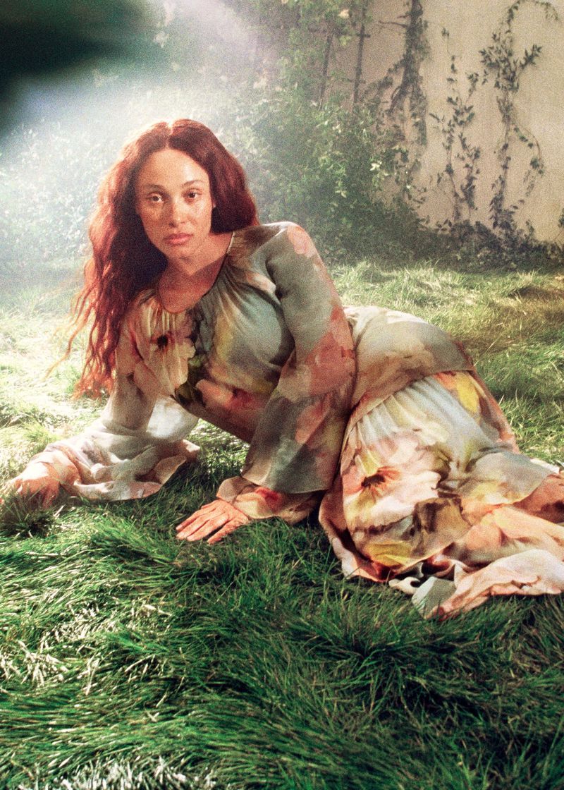 A woman with auburn hair reclines in a floral dress on green grass in a sunlit outdoor setting, creating a natural and artistic portrait.