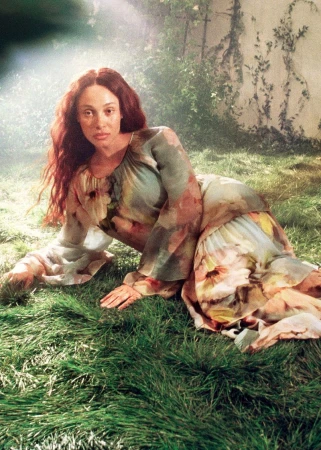 A woman with auburn hair reclines in a floral dress on green grass in a sunlit outdoor setting, creating a natural and artistic portrait.
