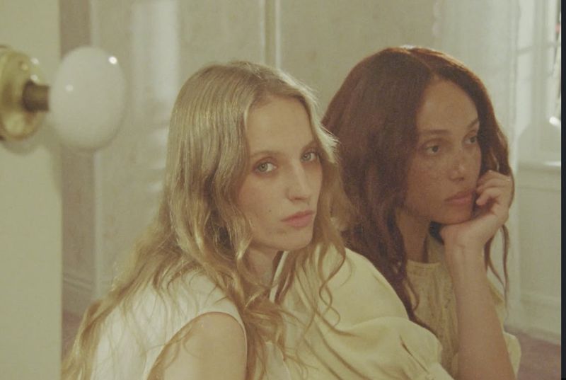 Portrait of two adult women in a close-up shot, one with light hair and the other with auburn hair, in an indoor studio setting.
