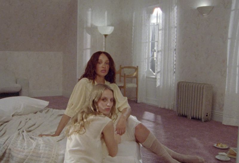 Two women in vintage-style dresses sit on a bed in a room with patterned wallpaper, sheer curtains, and a radiator in soft lighting.