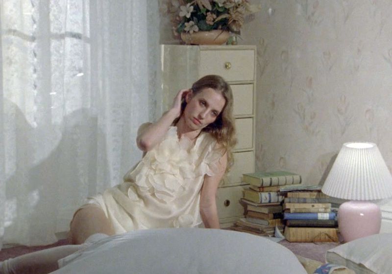 Woman in a pale yellow dress sits on a bed, resting her head on her hand, in a bedroom with patterned wallpaper and white window.
