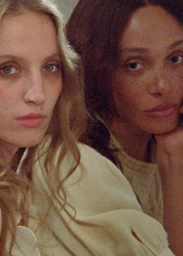 Close-up portrait of two female models with long hair wearing cream-colored textured garments in soft, grainy lighting.