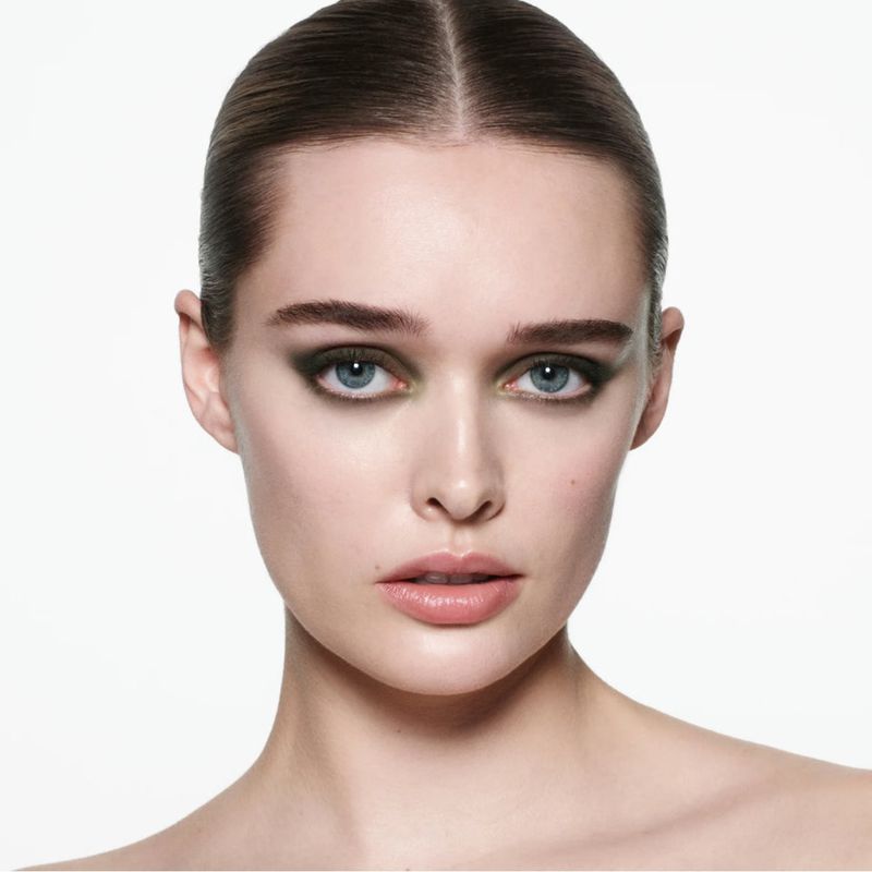 Close-up studio shot of a young adult woman with olive green smokey eye makeup and slicked-back hair, set against a bright white background.