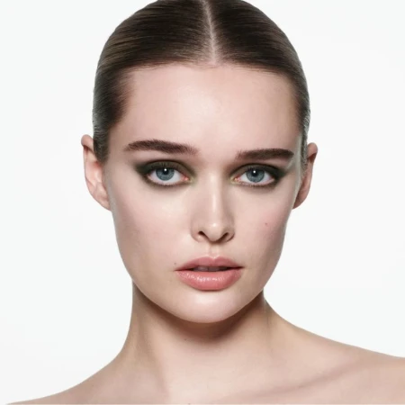 Close-up studio shot of a young adult woman with olive green smokey eye makeup and slicked-back hair, set against a bright white background.