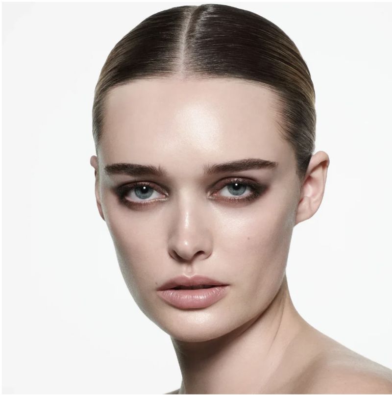 Close-up beauty portrait of an adult female with blue eyes, slicked-back brown hair, and dramatic black smoky eye makeup on a white backdrop.