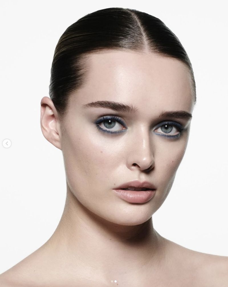 Close-up studio shot of an adult female with blue eye makeup, neutral-toned skin, and slicked-back dark hair against a bright white background.