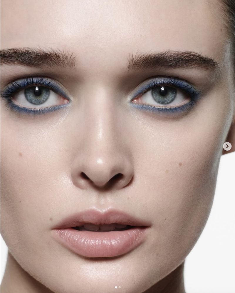 Close-up beauty shot of a fair-skinned adult female's face with blue eyeliner on the upper and lower eyelids, against a white backdrop.
