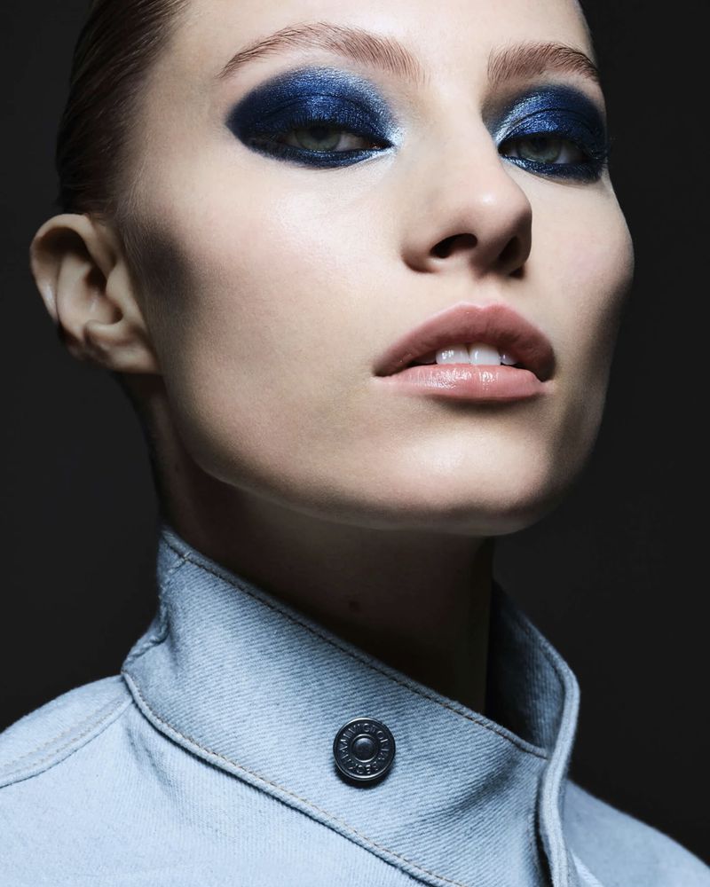 Close-up of a fair-skinned adult female with blue eyeshadow wearing a light blue denim jacket against a dark background.