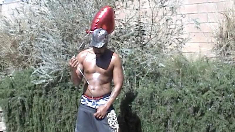Shirtless man holding a red balloon in an outdoor setting with green foliage and a brick wall in the background during the daytime.