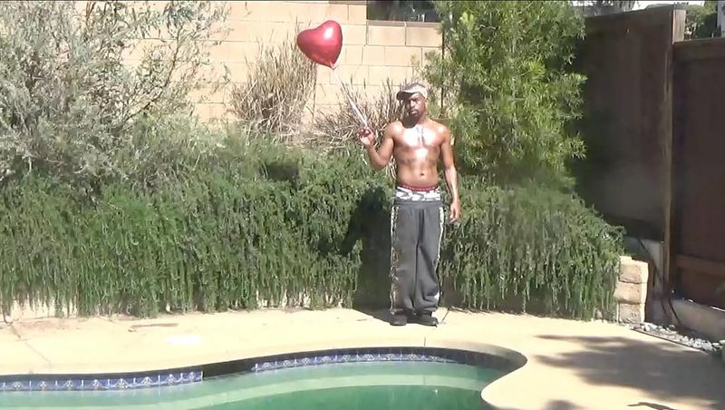 A man stands shirtless near a pool, holding a red heart balloon. He wears sweatpants and a hat in an outdoor setting with greenery.