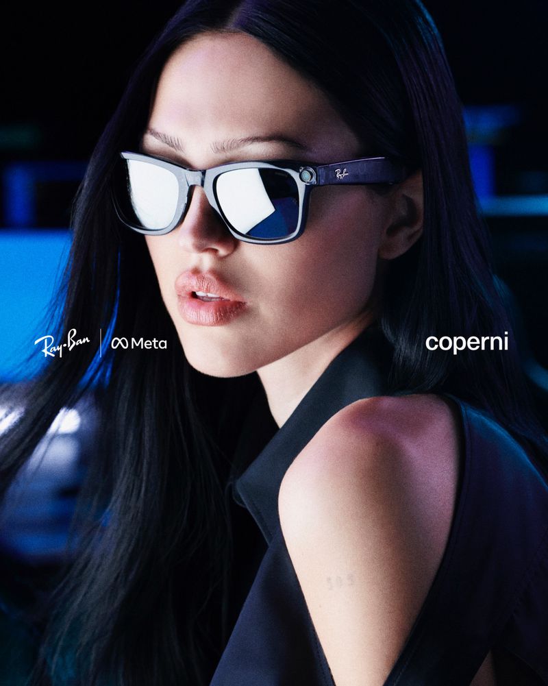 Adult woman with long black hair poses indoors wearing Ray-Ban Meta smart glasses in a fashion shot with product and brand logos.