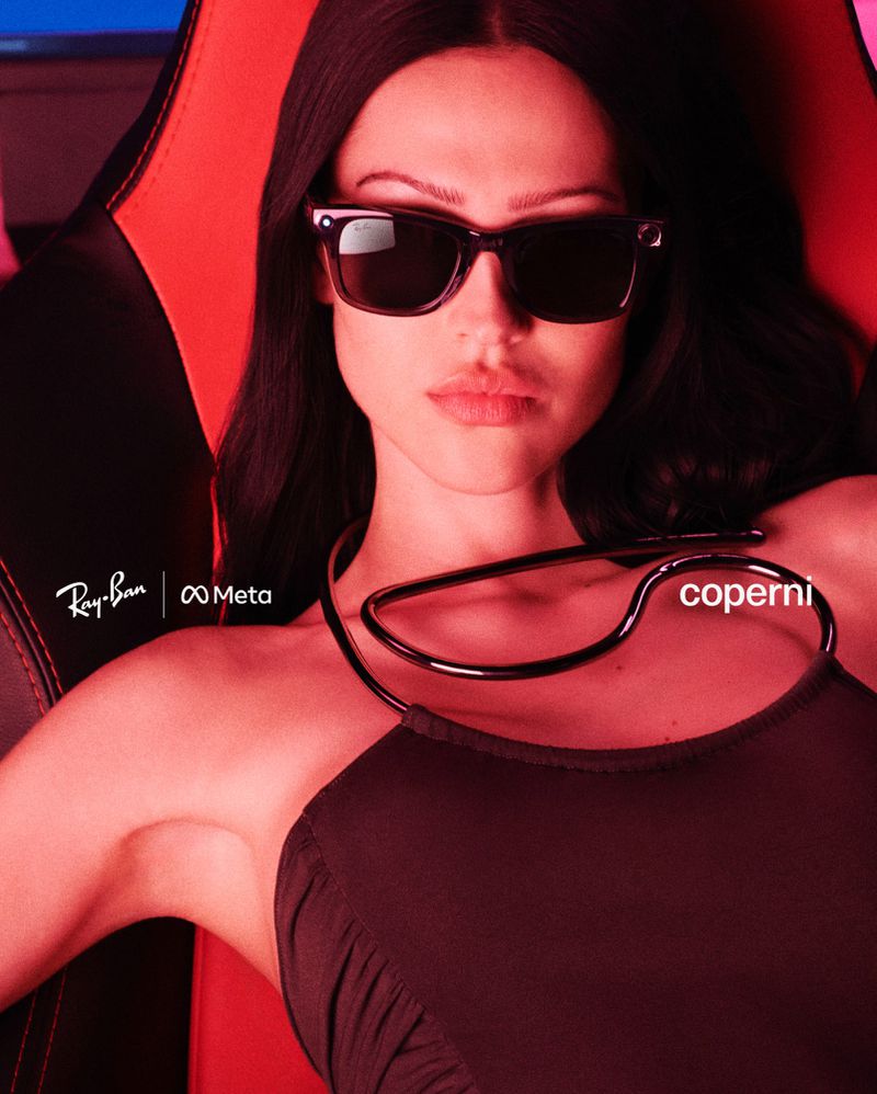 Indoor shot of a stylish adult female wearing sunglasses and a black dress, seated in a red and black gaming chair against a blue screen.