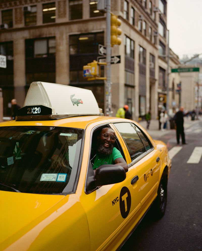 A yellow taxi cab in New York City shows a smiling African-American male driver leaning out the window with city buildings in the background.