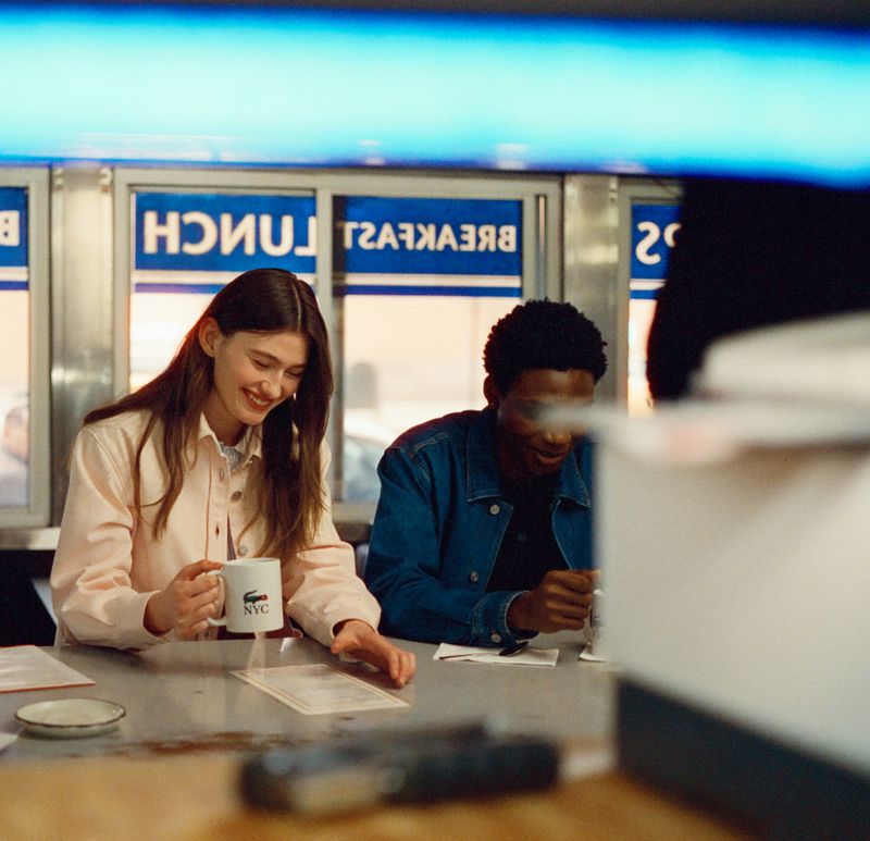 A smiling woman holding a mug with the Lacoste logo sits next to a man in a diner setting, both looking at a menu.