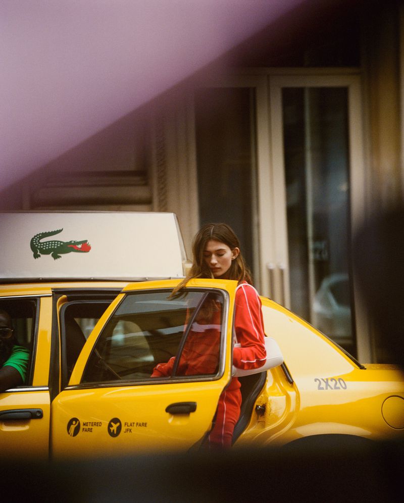 Woman exiting yellow taxi cab in an urban setting. She is wearing a red tracksuit. The taxi has an advertisement on top.