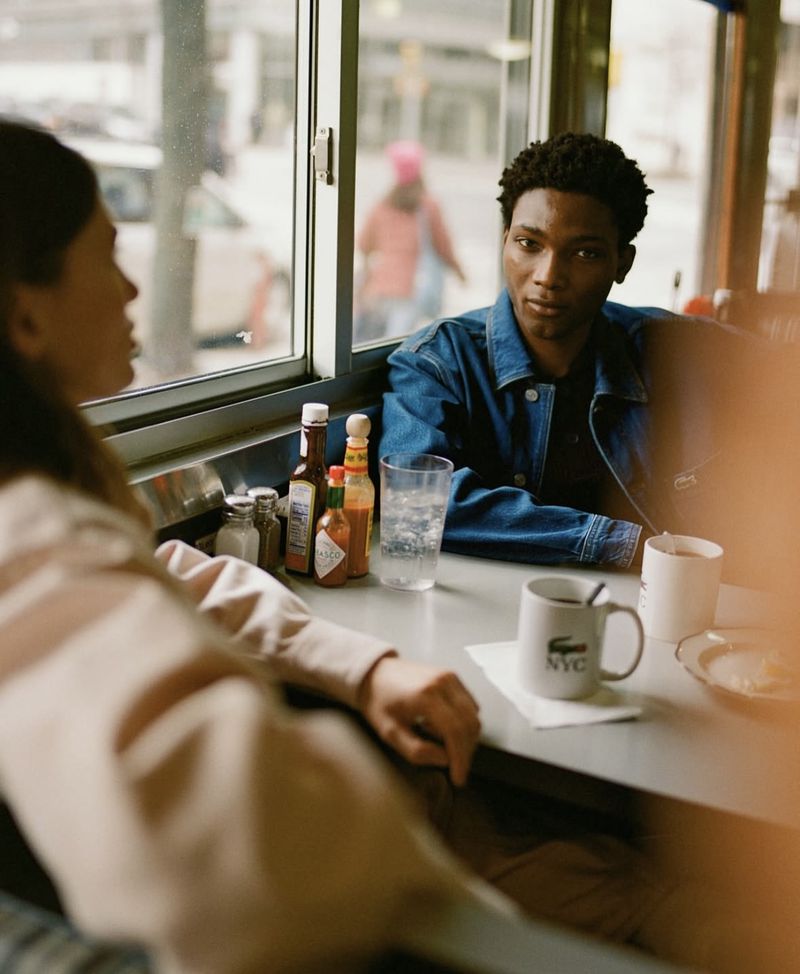 An African American man sits in a diner across from a woman, conversing over coffee near a window with a city street view.