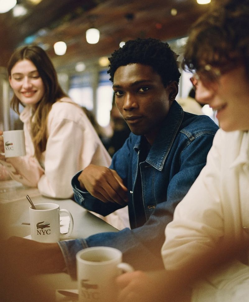 Three people, including an African-American man, seated at a counter with white mugs, wearing casual attire in an indoor setting.