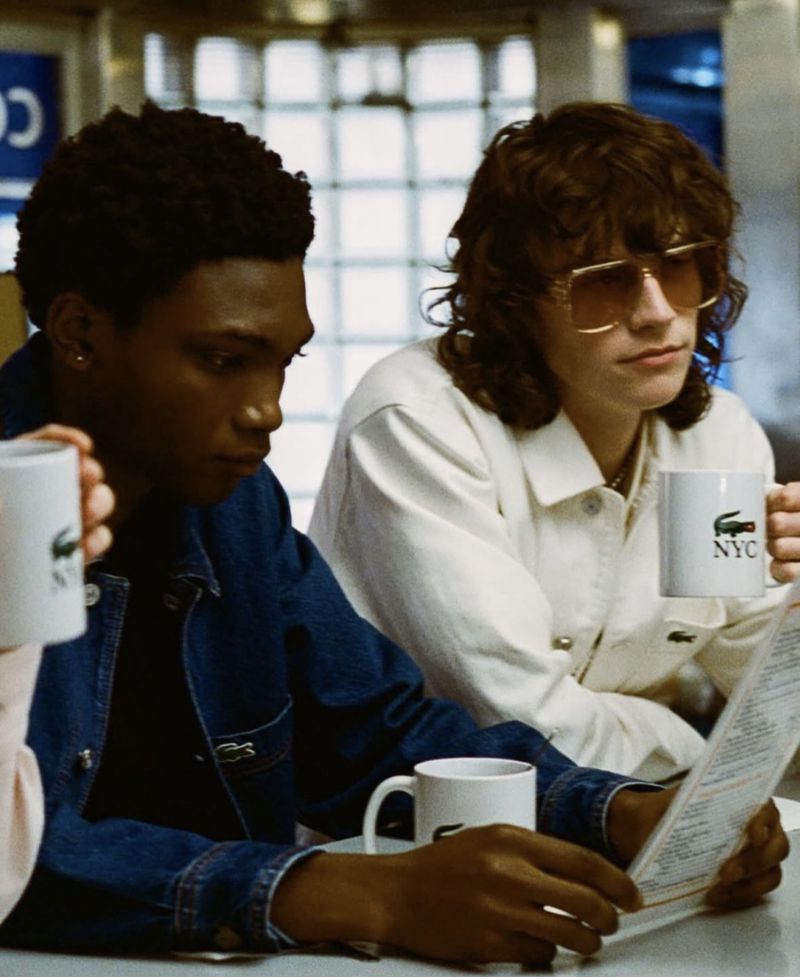 Two young adults in an indoor diner setting, wearing stylish jackets and holding branded mugs, focusing on a menu in a close-up shot.