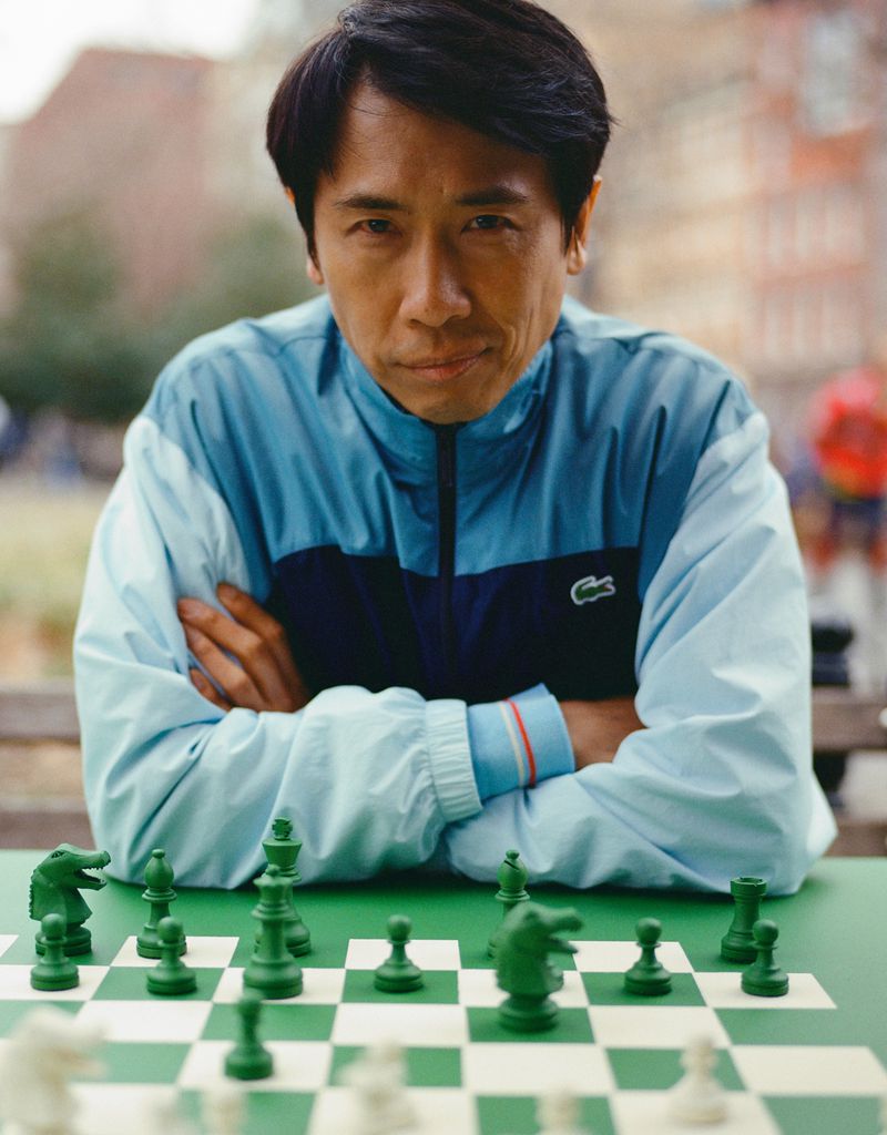 Portrait of an Asian adult male in a blue tracksuit seated at an outdoor table ready to play a game of chess with green pieces.