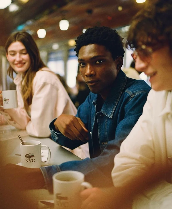 Three models seated at a diner counter holding branded mugs during the Lacoste 5th Avenue campaign.