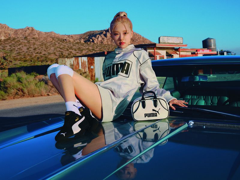 Woman in sporty outfit poses on a vintage car in a desert landscape with mountains and industrial building during daytime, wearing Puma brand.