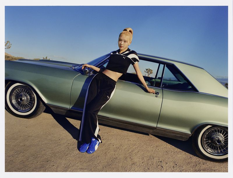 Adult woman in Puma athletic wear poses against a light-green vintage car in a desert landscape under a clear blue sky.