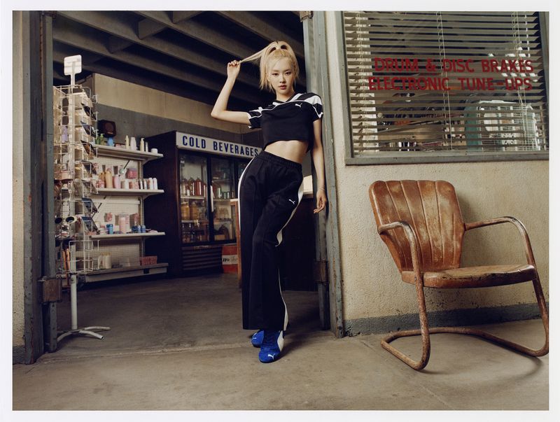 A woman in athletic wear poses in front of a garage-like space with beverage cooler and vending machine, with chair and lettering in the scene.