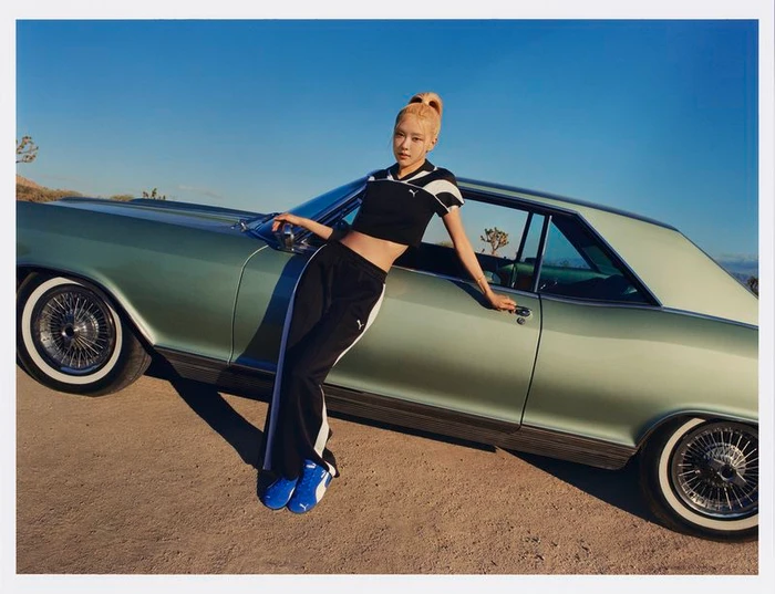 Rosé Park leaning against a vintage green car wearing a black athletic crop top and track pants under a clear blue sky