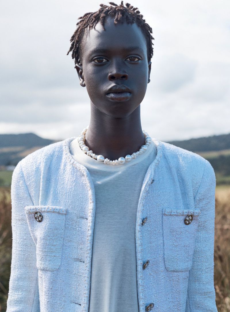 Close-up of an adult with an afro hairstyle, wearing a tweed jacket and pearl necklace in an outdoor field with a cloudy sky.