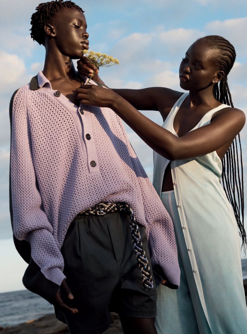 Two dark-skinned women outdoors. One woman holds small yellow flowers near the other woman's chest, who wears a lilac sweater and dark shorts.