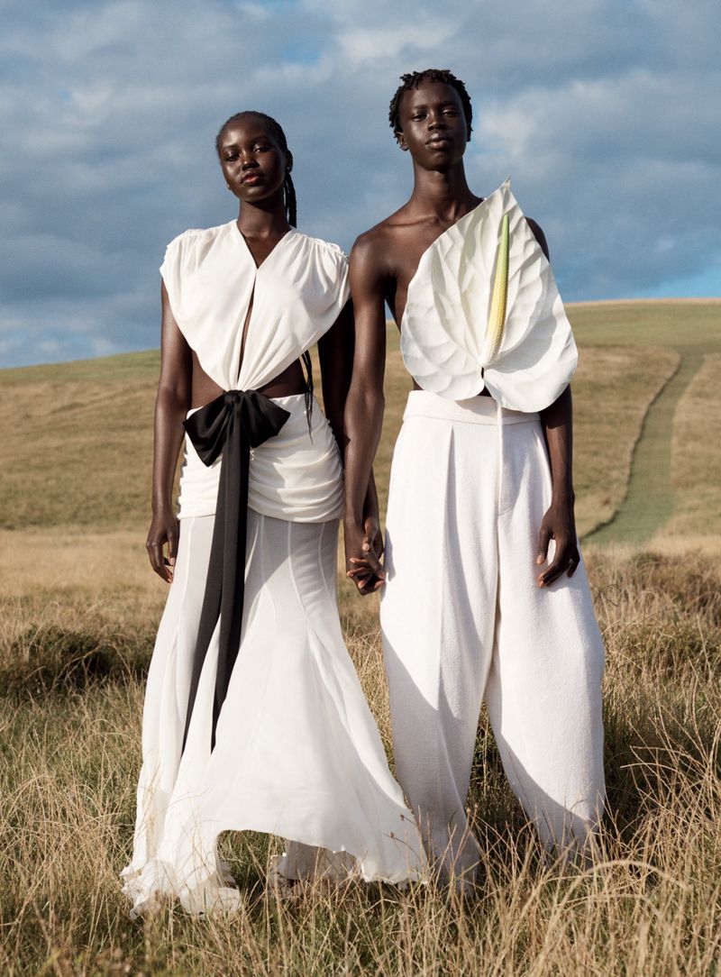 Two models in draped white and black couture stand in a field against rolling hills and cloudy sky, holding hands, fashion shoot.