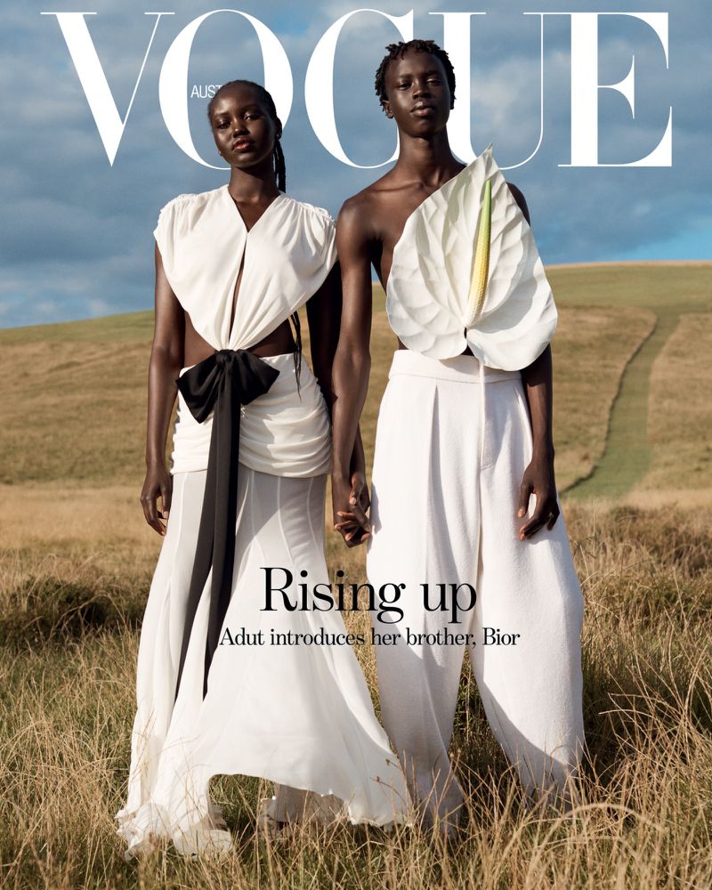 Vogue cover featuring two African models in flowing white outfits, holding hands in a grassy field under a cloudy sky in an outdoor setting.