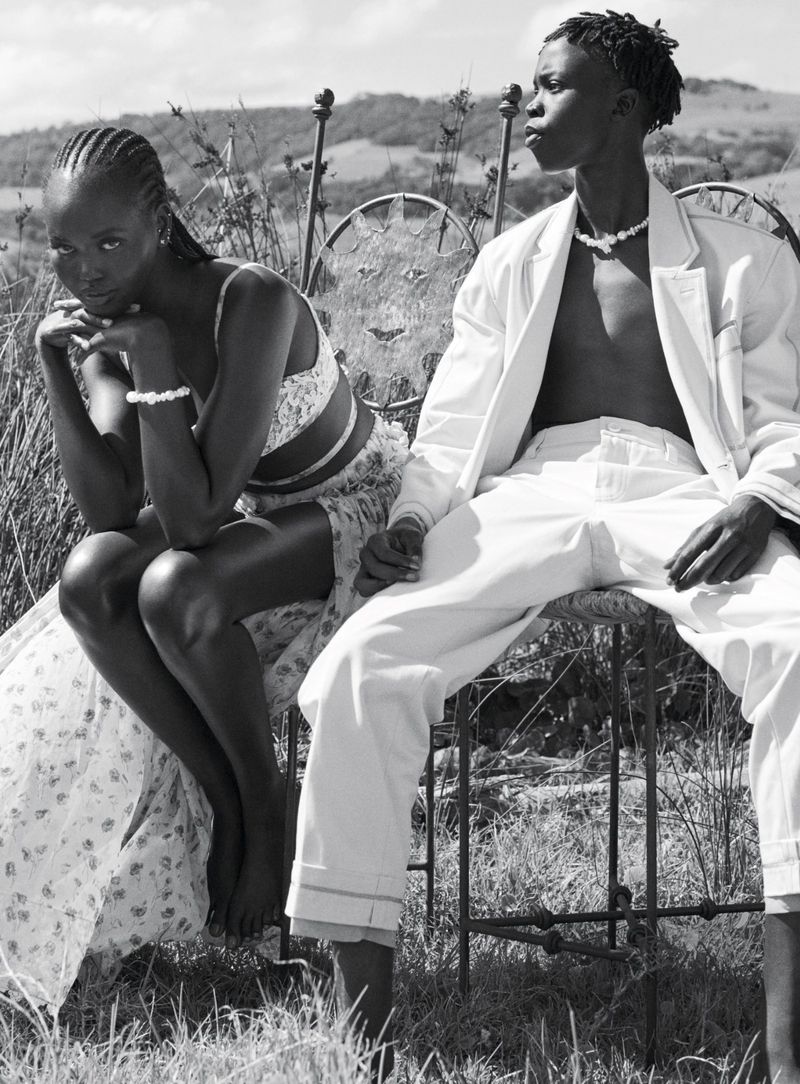 Monochrome portrait of a young African woman in floral top and a young man in a white suit, sitting on an ornate metal bench outdoors.