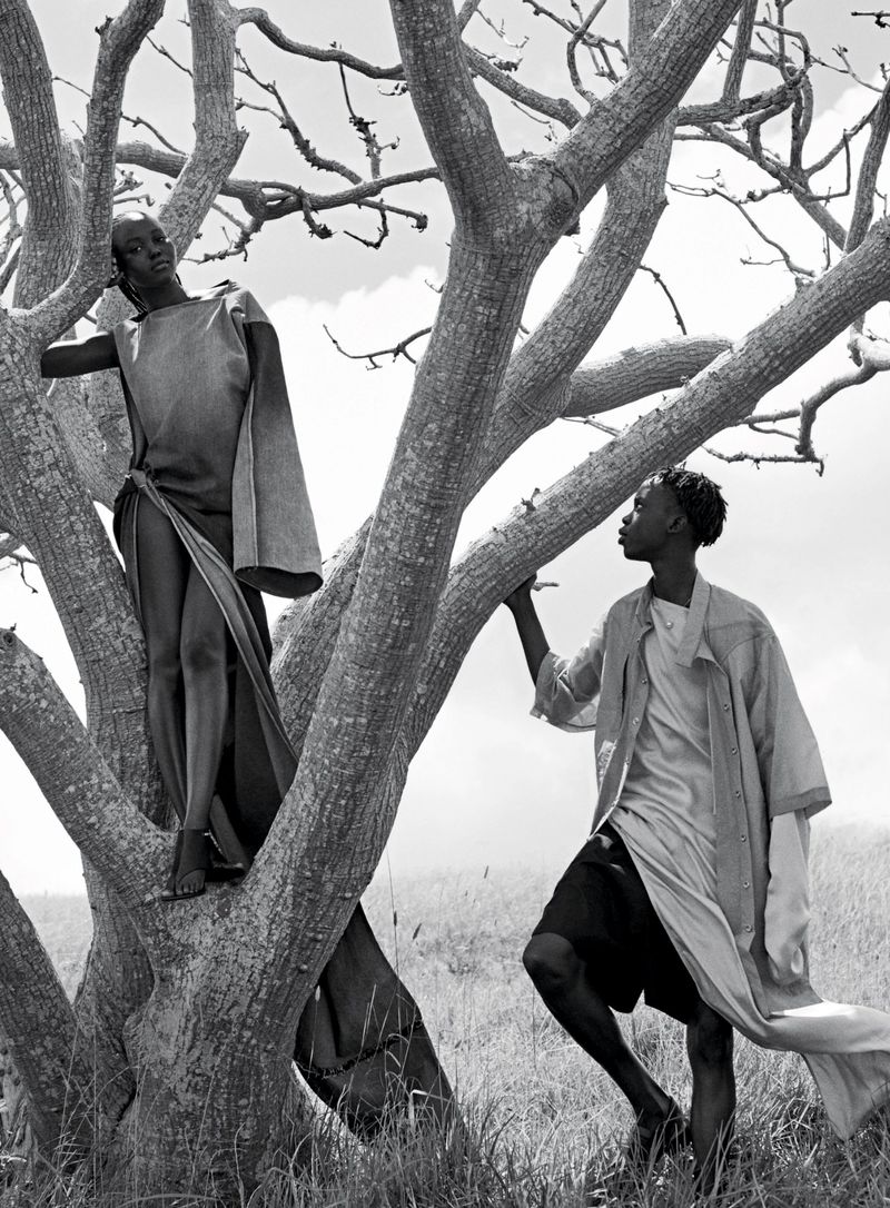 Black and white fashion photo of two dark-skinned adults in flowing robes standing in a bare tree on a grassy hill under a cloudy sky.