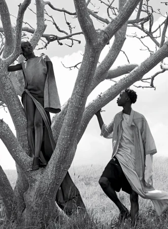 Black and white fashion photo of two dark-skinned adults in flowing robes standing in a bare tree on a grassy hill under a cloudy sky.